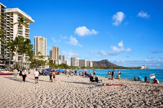 Plaża Waikiki w Honolulu z piaskiem, ludźmi, palmami, wieżowcami i oceanem.